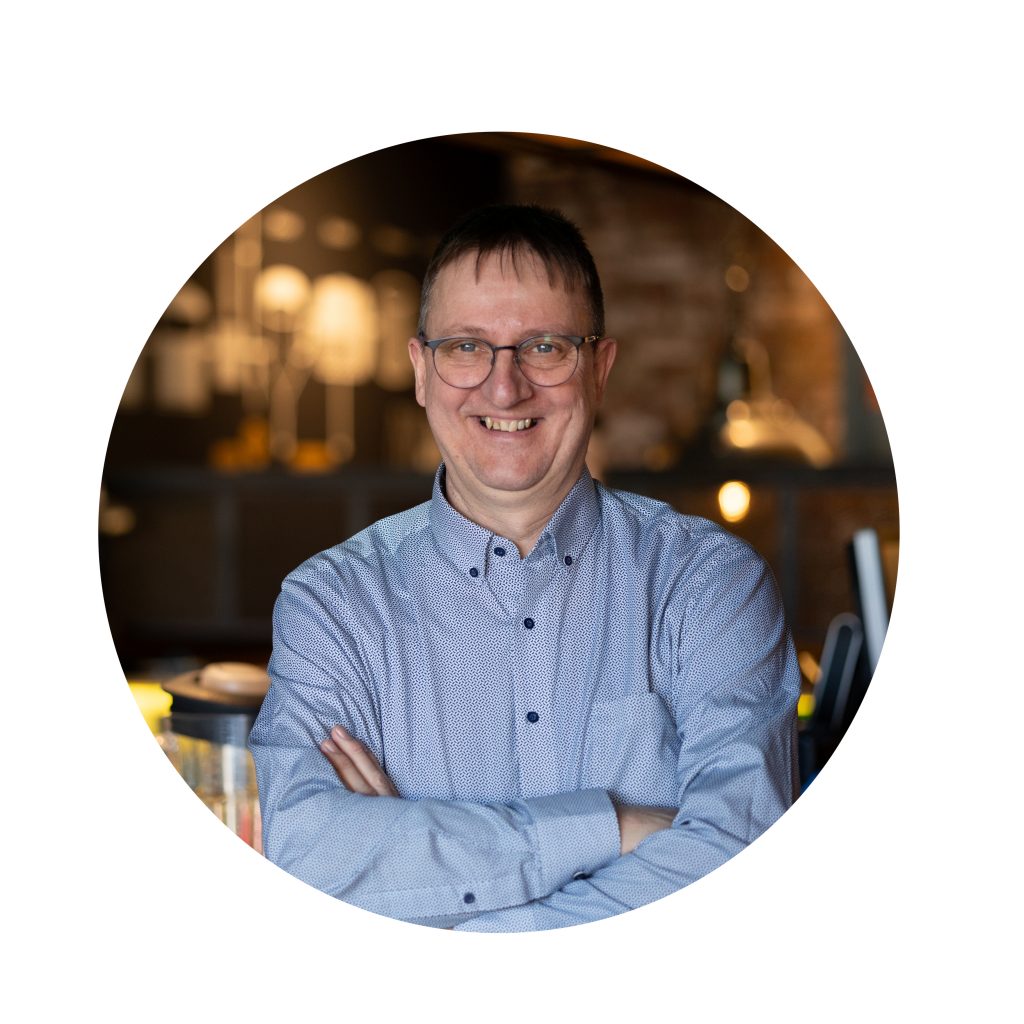 Mann mit hellblauem Hemd steht mit verschränkten Armen vor unscharfer, warm beleuchteter Bar, kreisförmig freigestelltes Businessportrait für Gastronomie