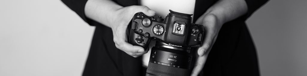 Fotografin aus Köln hält schwarze Kamera mit großem Objektiv in beiden Händen, schwarz-weiße Studioaufnahme