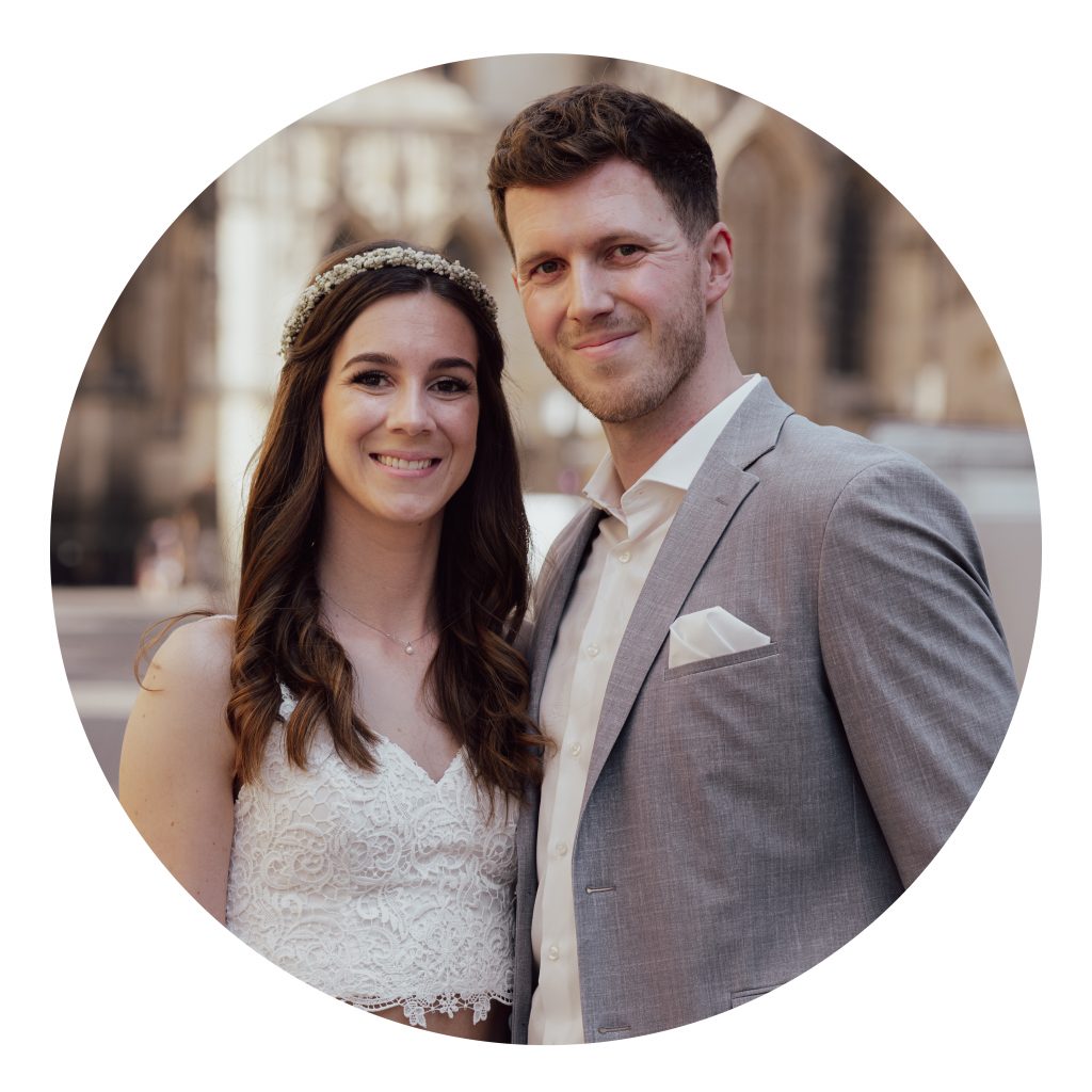 Brautpaar steht eng nebeneinander vor urbanem Hintergrund, sie im Spitzenkleid mit Haarband, er im hellen Anzug, kreisförmig freigestelltes Hochzeitsfoto