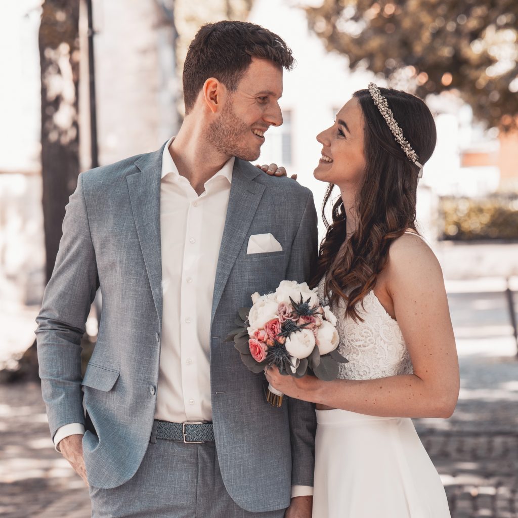 Brautpaar im Freien in Köln, sie im weißen Spitzenkleid mit Brautstrauß, er im hellen Anzug, halten sich eng im Arm